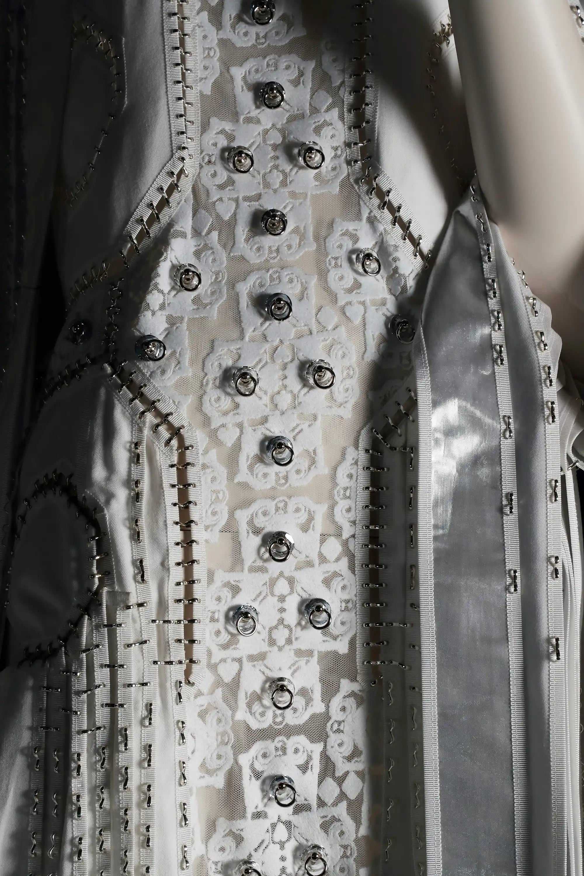 Close-up detail of the front middle section of The Florentine Dress. Digitally designed and embroidered white lace depicts monster faces, with crystals attached to their mouths with door-knocker style embellishments. The creation is seamless and the angles of the front embroidered section attach to larger eye style cuts and layers, crafted in white and silver viscose and silk. The dress is seamless - connected with sculpted silver links creating a highly detailed and decorative presence.