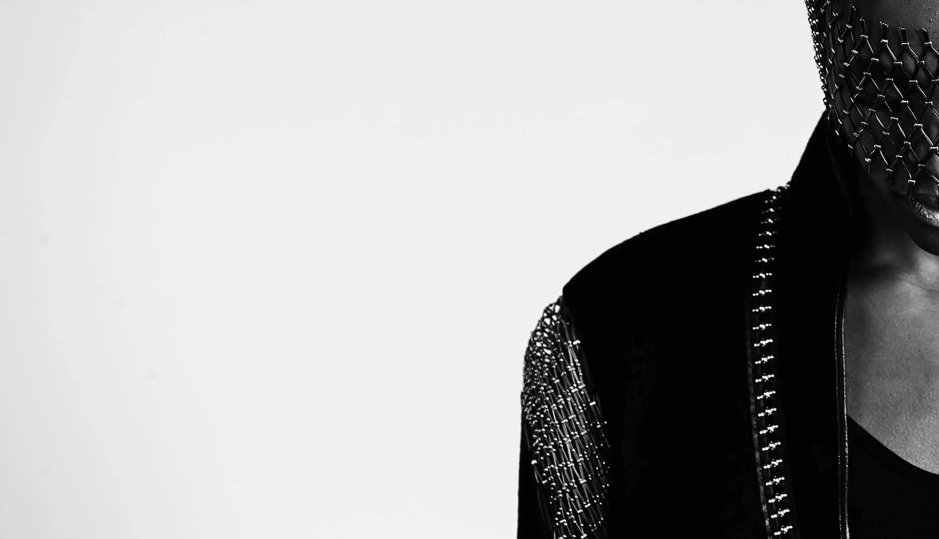 The Rila Coat and mask from Lorn Jean's "Diadem" collection, styled on a model in a portrait style with the model looking down, their face covered by a lattice jewellery piece. The image is cropped to show only the left side of the model, in black and white, creating a powerful depiction. The shoulder detail of the coat is fully constructed with integrated jewellery and the collar is connected with a line of sculpted silver links.